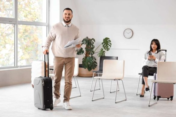 Un homme souriant avec une valise cabine attend dans une salle lumineuse, prêt à voyager avec un petit budget.