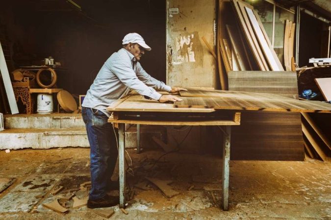 Un artisan menuisier en casquette blanche ajuste des panneaux en bois sur son établi dans un atelier.