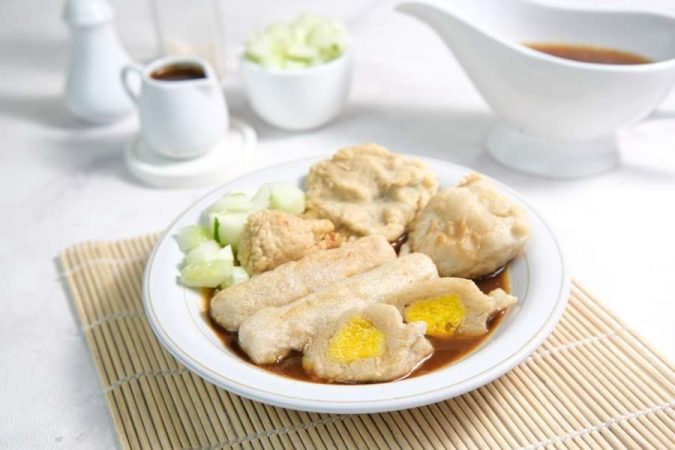Une assiette de pempek indonésien avec de la sauce cuko et des concombres sur un set de table en bambou, invitant à un voyage culinaire exotique.