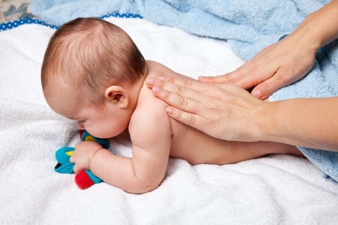 Un bébé souriant allongé sur le dos est massé sur le ventre par les mains d'un adulte.