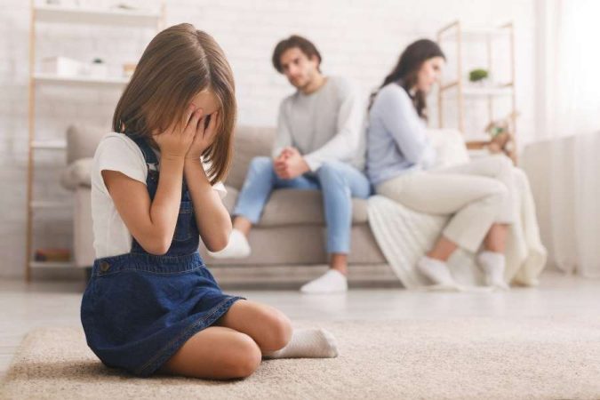 Une petite fille pleure, cachant son visage, tandis que ses parents se disputent en arrière-plan, illustrant la détresse liée à une séparation.